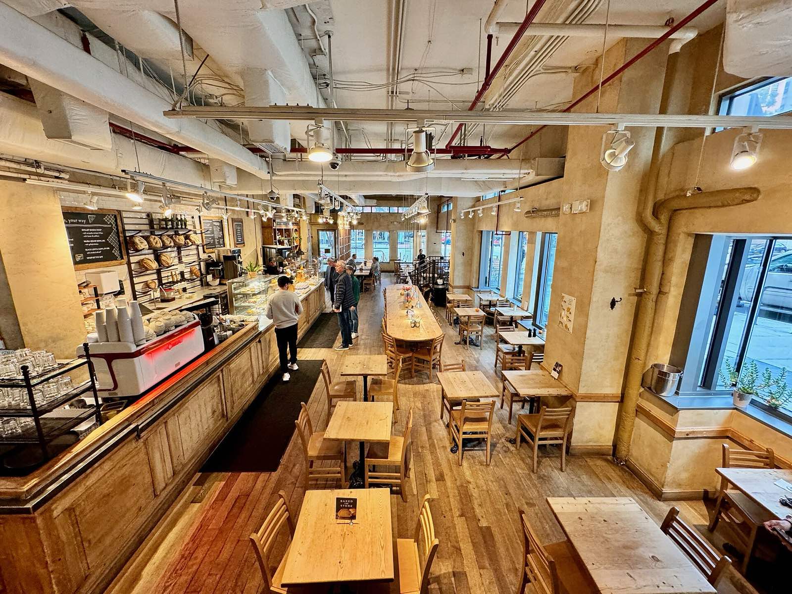 A long, sunlit cafe room with wooden tables, a pastry counter and an espresso bar